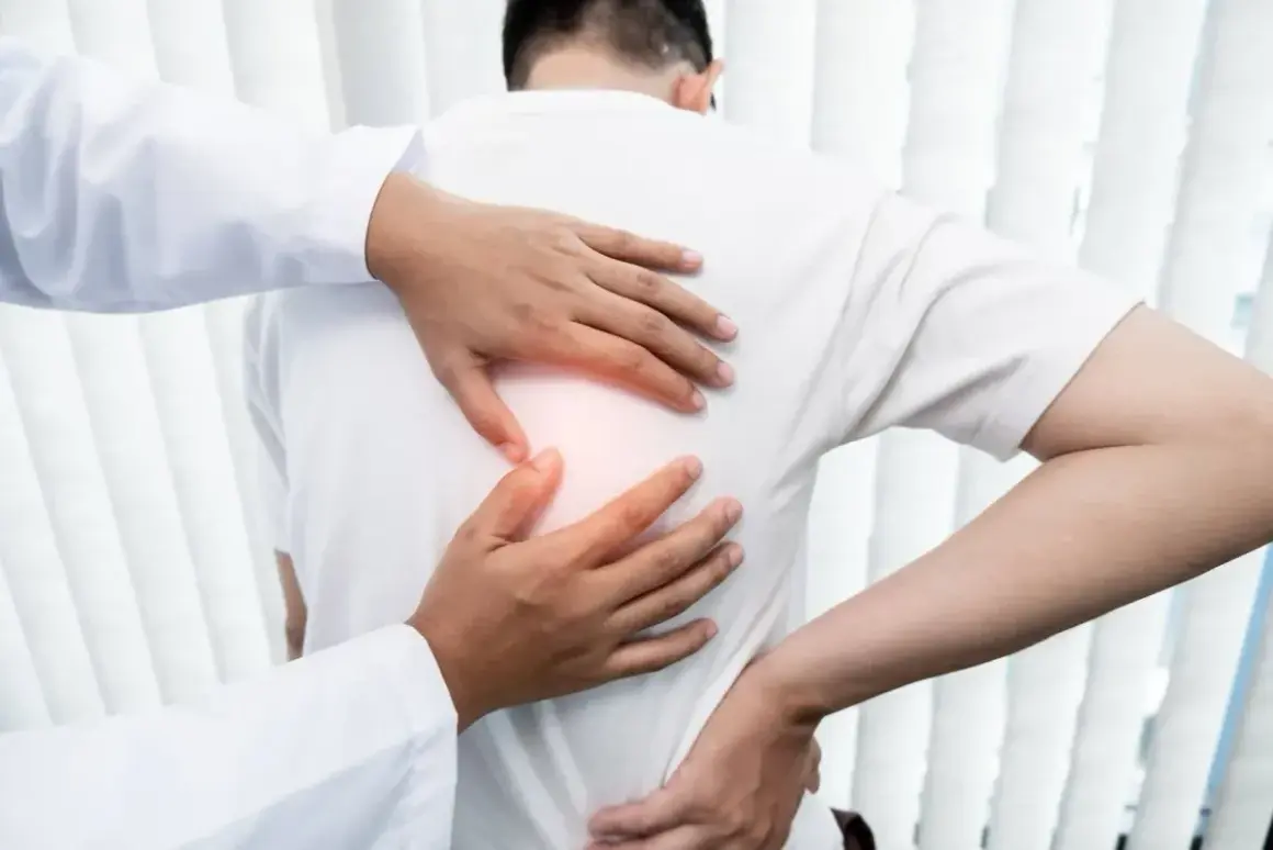 Lekarz bada pacjenta z bólem pod lewą łopatką. Dłonie terapeuty delikatnie dotykają pleców, wskazując na źródło dyskomfortu.